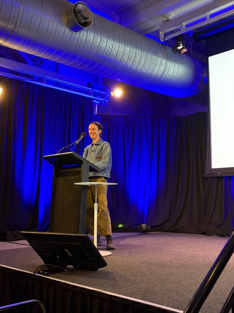 A person standing at a podium in front of a screen, smiling and engaging with the audience during a presentation in a well-lit venue.